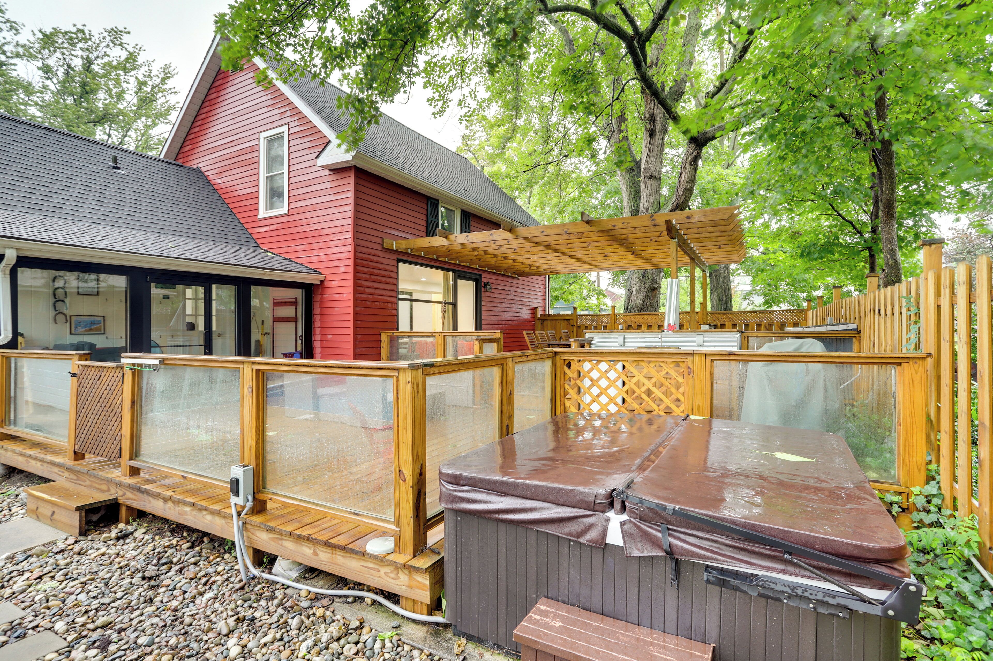 Un jacuzzi con un deck de madera, una casa roja y un área cubierta con una pérgola de madera.