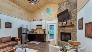 A cozy living room with a fireplace, wooden ceiling, and a stone wall. There is a kitchen in the background with white cabinets and a refrigerator. - Blue Ridge Mountainside Cottage w/ Fire Pit! (Epworth)