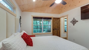 A bedroom with a large bed, a ceiling fan, a window with a view of trees, and a wooden door. - Blue Ridge Mountainside Cottage w/ Fire Pit! (Epworth)