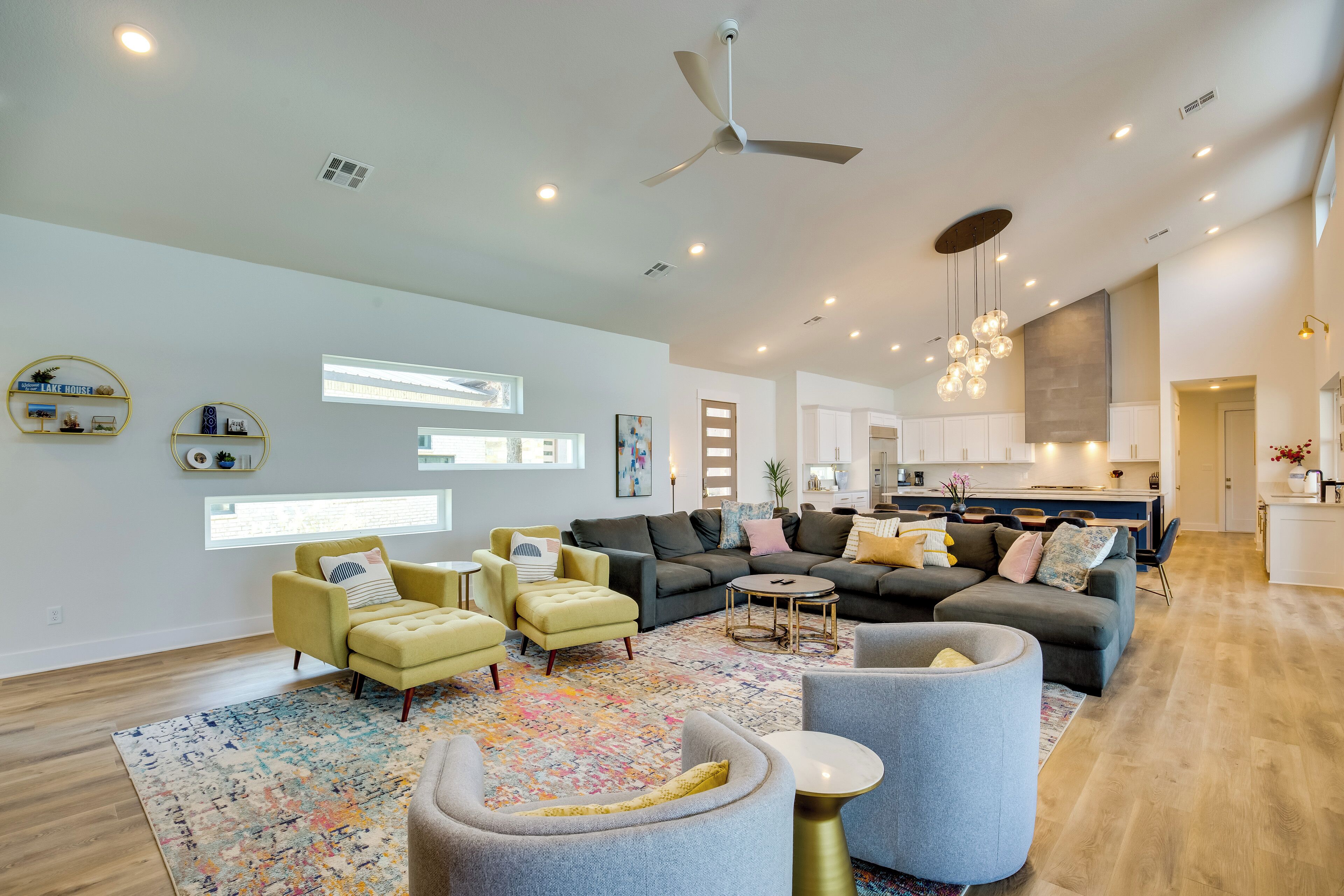 Un salón espacioso con un sofá grande, sillones y una mesa de centro. Se observa una cocina al fondo con armarios blancos y una mesa de comedor.