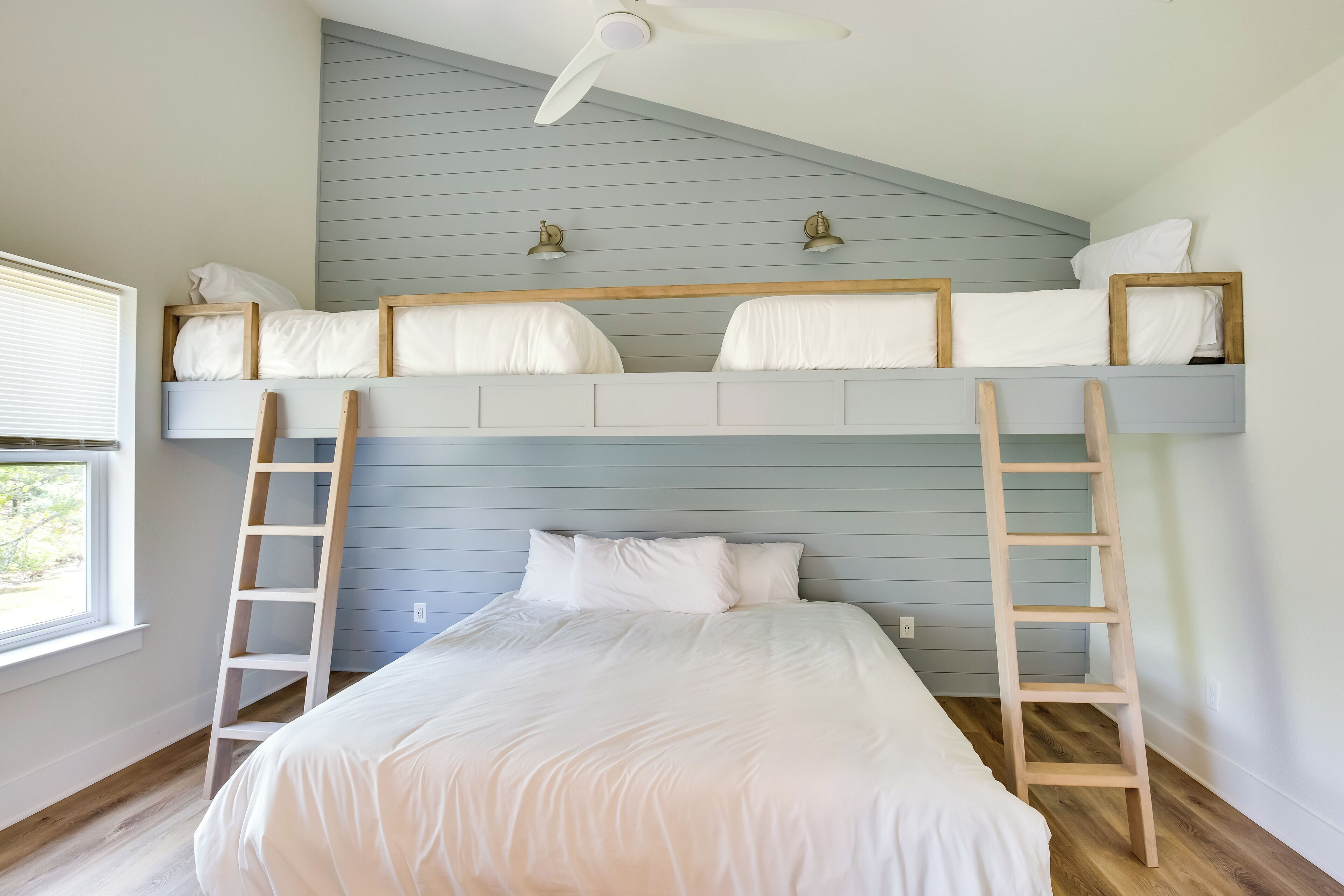 A bunk bed room with a ceiling fan, wooden ladders, and a window with blinds.