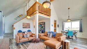 Un salón espacioso con techo alto, un balcón de madera y una zona de comedor con sillas azules.