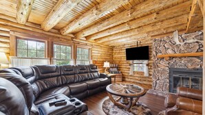 A log cabin interior with a leather sectional sofa, a stone fireplace, and a mounted television.