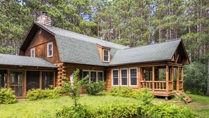 A log cabin with a porch, a pond, and a forest backdrop.