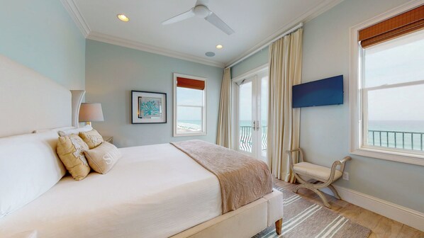 A hotel room with a bed, a ceiling fan, a window with curtains, a framed picture, and a TV mounted on the wall. - Dos Casas - Gulf-front Home with 2 Martini Pools and Beach Service (Destin)