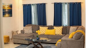 Un salón con un sofá gris, cojines estampados en amarillo y blanco, una mesita redonda y un jarrón azul con flores.
