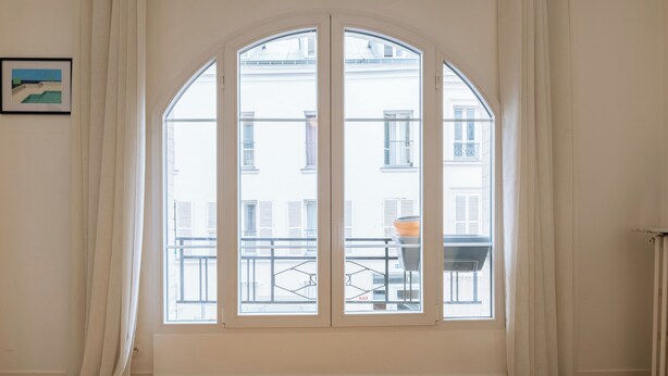 Una habitación con un amplio ventanal de arco, cortinas blancas, un cuadro en la pared y vistas a edificios y un balcón.