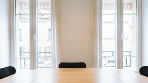 Un salón de conferencias con una larga mesa y dos sillas, ventanas amplias con cortinas blancas y vistas a edificios exteriores.