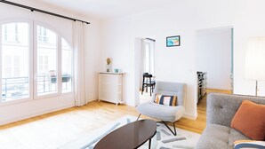 Un salón luminoso con ventanas amplias, un radiador, una zona de comedor y una cocina visible a través de una puerta abierta.