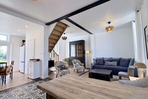 Un vaste espace de vie comprenant une table basse en bois, un canapé et des fauteuils. Un escalier mène à l’étage supérieur.