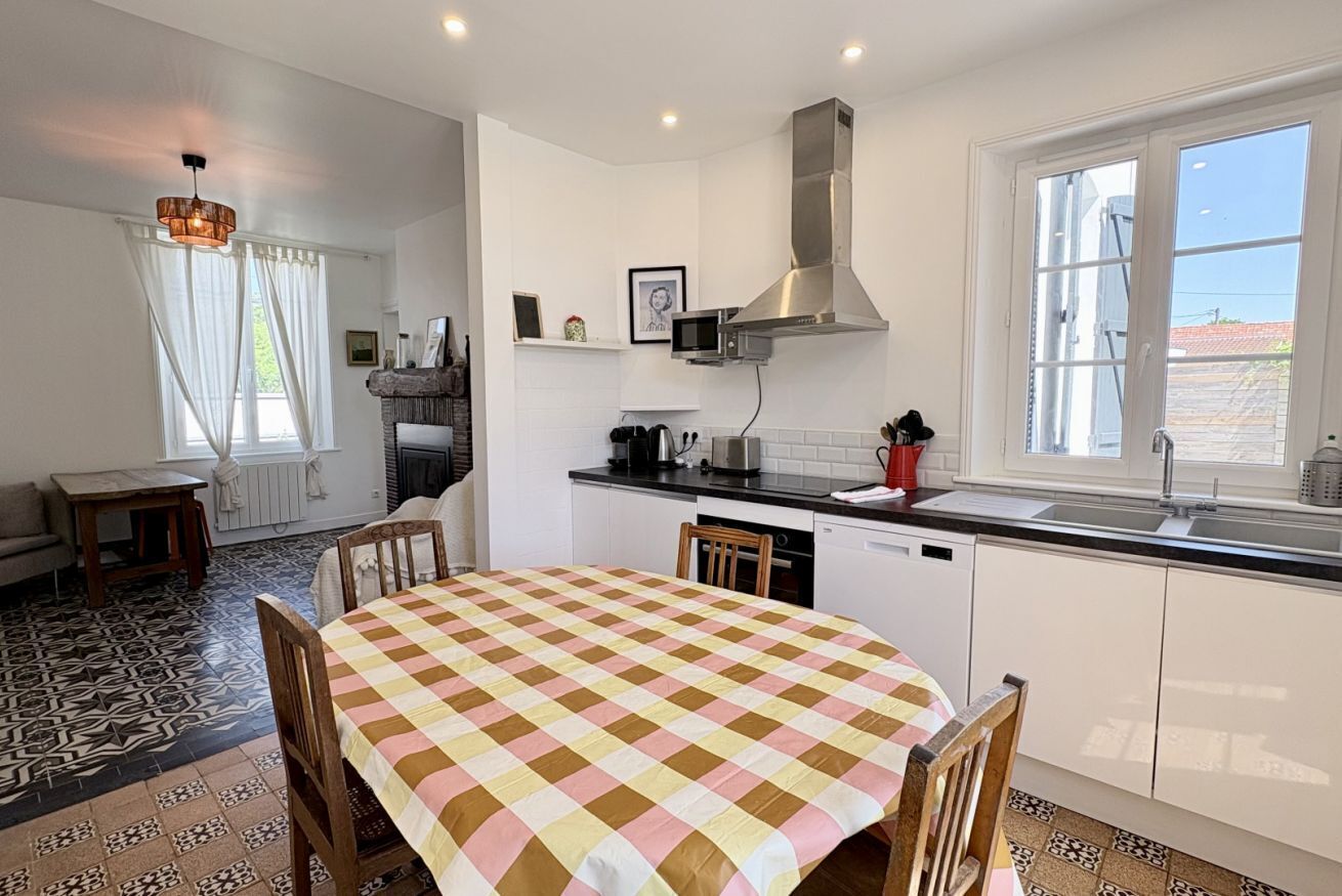 Une cuisine moderne avec des armoires blanches, une hotte de cuisinière en acier inoxydable, une table à manger recouverte d’une nappe à carreaux et une grande fenêtre.