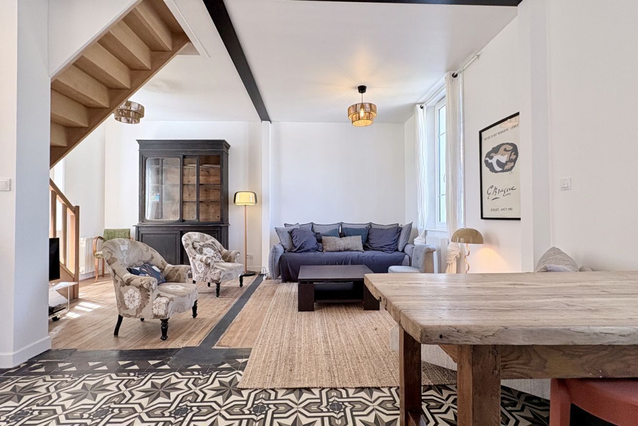 Un salon comprenant un canapé, des fauteuils, une table basse, un tapis à motifs, une table à manger en bois et un escalier.
