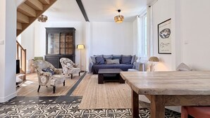 A living room with a sofa, armchairs, a coffee table, a patterned rug, a wooden dining table, and a staircase.