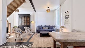 Un salon comprenant un canapé, des fauteuils, une table basse, un tapis à motifs, une table à manger en bois et un escalier.