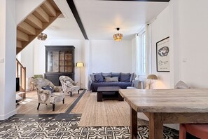 Un salon comprenant un canapé, des fauteuils, une table basse, un tapis à motifs, une table à manger en bois et un escalier.