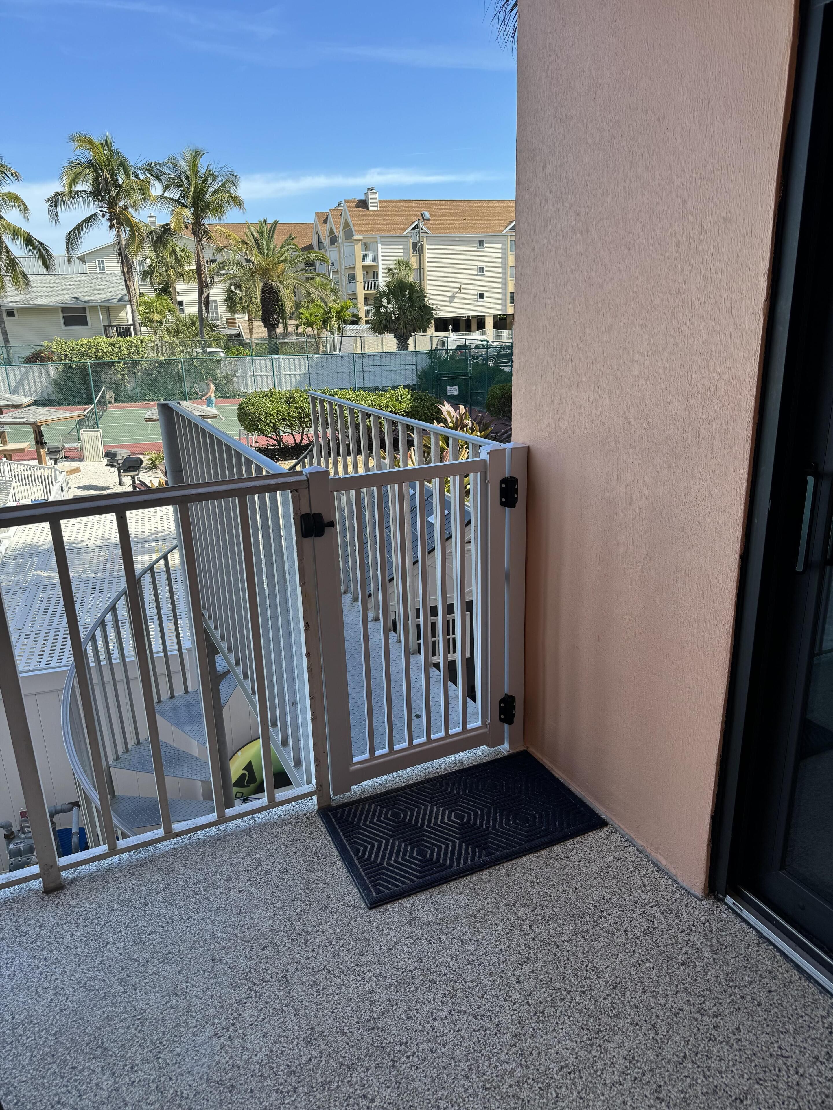 A balcony with a metal gate, a black mat, and a view of a tennis court and buildings in the background.
