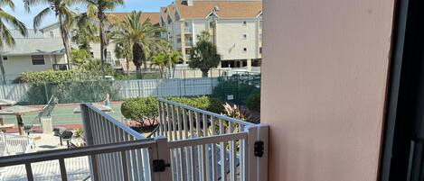 A balcony with a metal gate, a black mat, and a view of a tennis court and buildings in the background.