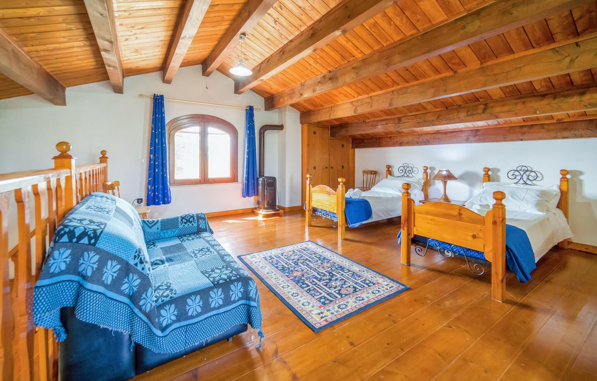 A cozy room with wooden beams, two beds with wooden frames, a sofa with a patterned blanket, and a large window with curtains.