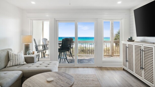 A spacious living room with a large sofa, a coffee table, and a TV on a cabinet. There is a sliding glass door leading to a balcony with a view of the ocean.