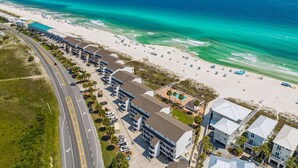 An aerial view of a coastal hotel with a beachside location, featuring a road with a parking lot, multiple buildings with balconies, and a swimming pool area.