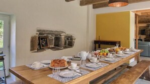 A long wooden dining table set for breakfast with a stone fireplace, wooden beams on the ceiling, and a yellow wall in the background. - Kerguntuil - Pleumeur Bodou (PLEUMEUR BODOU)