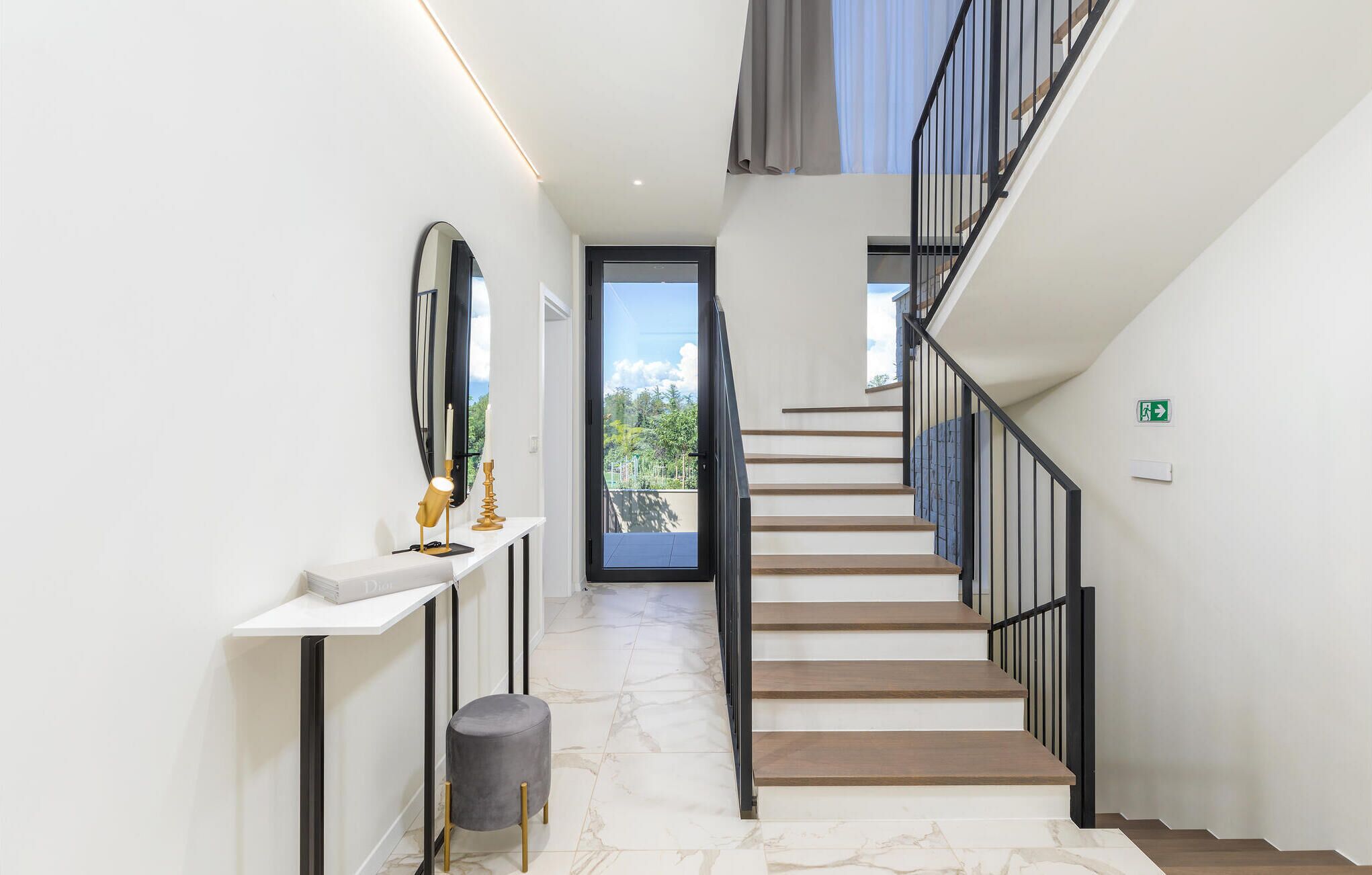 A modern staircase with a glass door leading to a balcony, a white console table with a lamp and candle, and a marble floor.