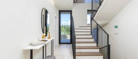 A modern staircase with a glass door leading to a balcony, a white console table with a lamp and candle, and a marble floor.