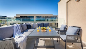 A rooftop terrace with a sofa set, a table with drinks and fruit, and a view of a modern building.
