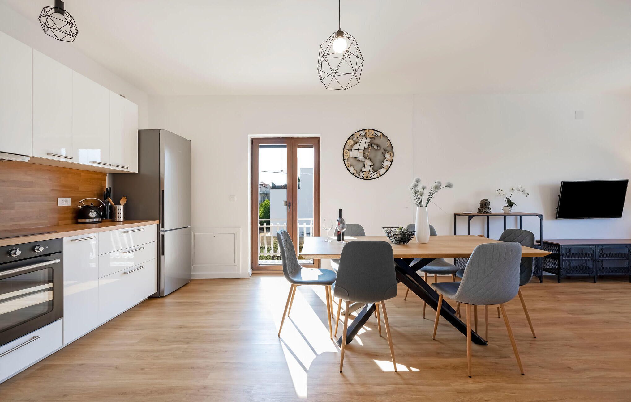 A modern kitchen with white cabinets, a wooden countertop, and a built-in oven. There is a dining area with a wooden table and chairs, a vase with flowers, and a flat-screen TV.