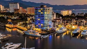 A waterfront hotel with multiple balconies, a marina with yachts, and a cityscape in the background.