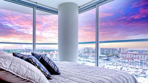 A bedroom with a large bed, multiple pillows, and a view of a marina and cityscape through large windows.