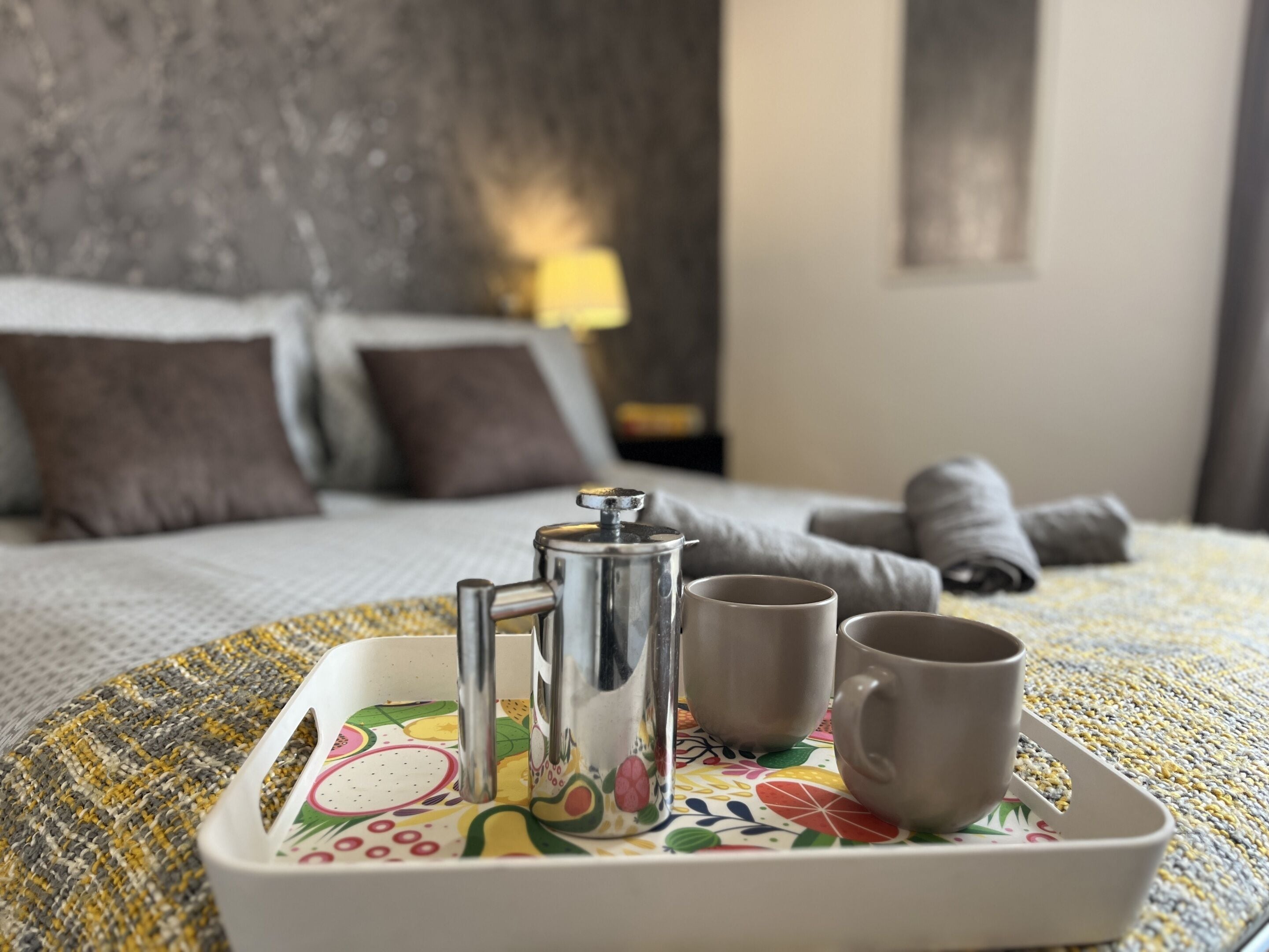 A neatly made breakfast tray with a coffee pot, two cups, and a teapot on a bed with pillows and a blanket.
