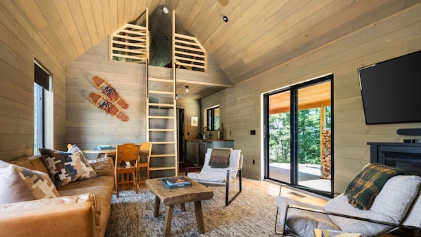 Un intérieur de chalet chaleureux avec une chambre à coucher située en mezzanine, un coin repas et un salon doté d’une cheminée.