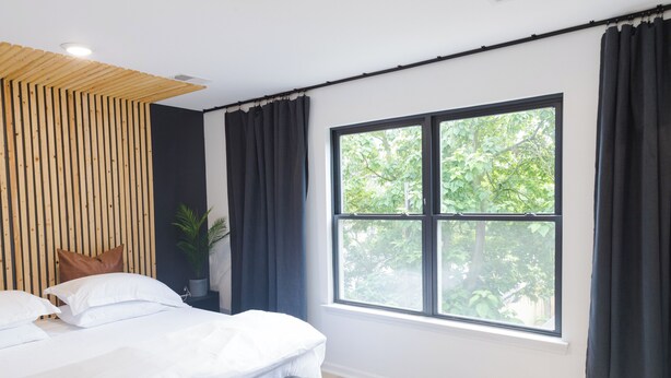Un dormitorio moderno con un ventanal grande, una cama con sábanas blancas, un cabecero oscuro y una pared de listones de madera.
