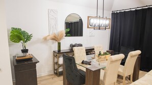 Un comedor con mesa de vidrio, sillas acolchadas, aparador de madera oscura, planta en maceta y espejo en la pared.
