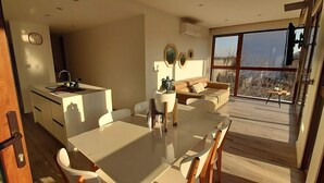 Una cocina moderna con una isla central, comedor con mobiliario blanco y un amplio ventanal corredera que da a una terraza con vistas al exterior.