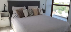 Una cama bien hecha con cabecera gris, una mesita de noche con lámpara y una ventana con vistas a edificios y árboles.