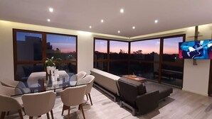 Una sala de estar moderna con una mesa de comedor de vidrio, asientos cómodos, un televisor de pantalla plana y grandes ventanas que ofrecen vistas a la ciudad.