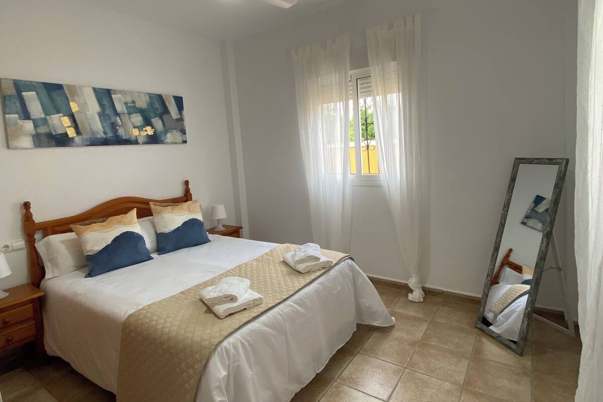 A neatly arranged bedroom with a bed, a bedside table, a mirror, and a window with curtains.