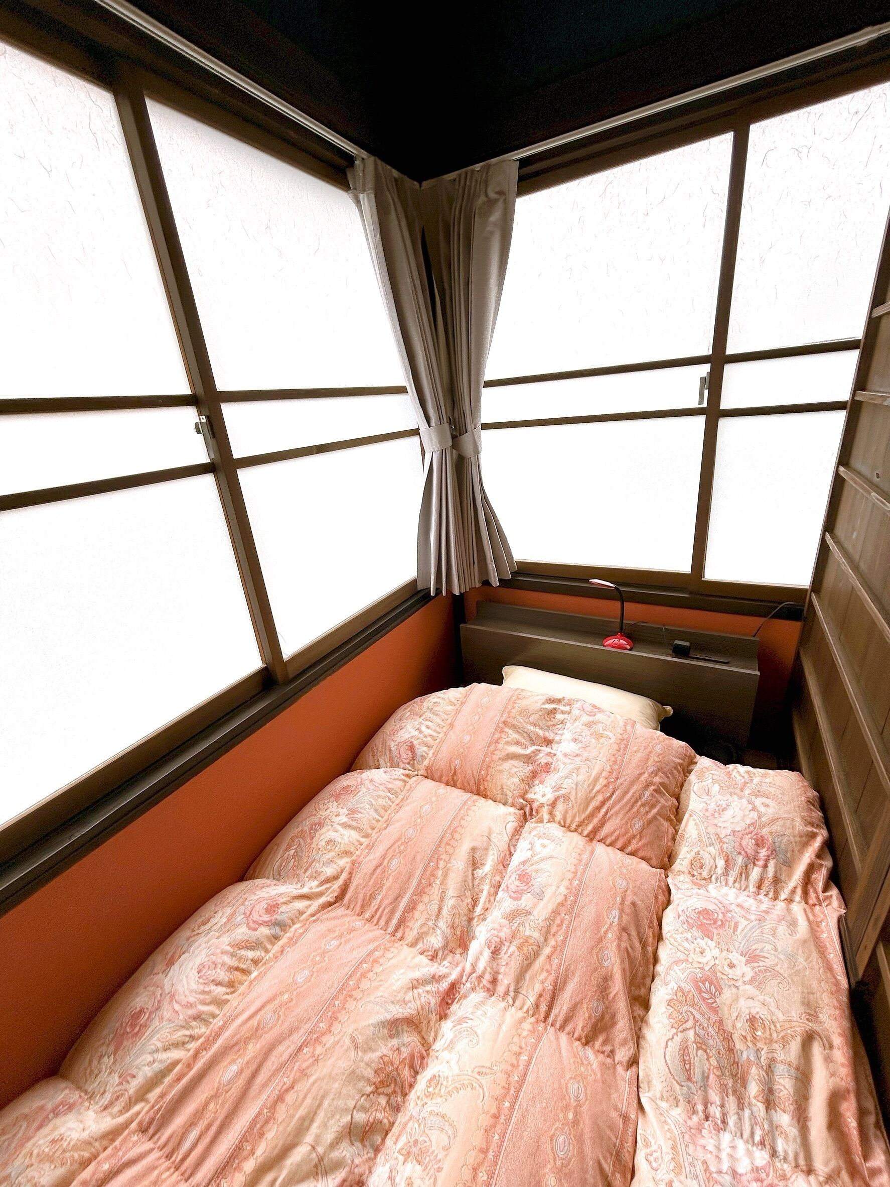 A bedroom with a bed, a nightstand, a window with curtains, and a view of the outside.