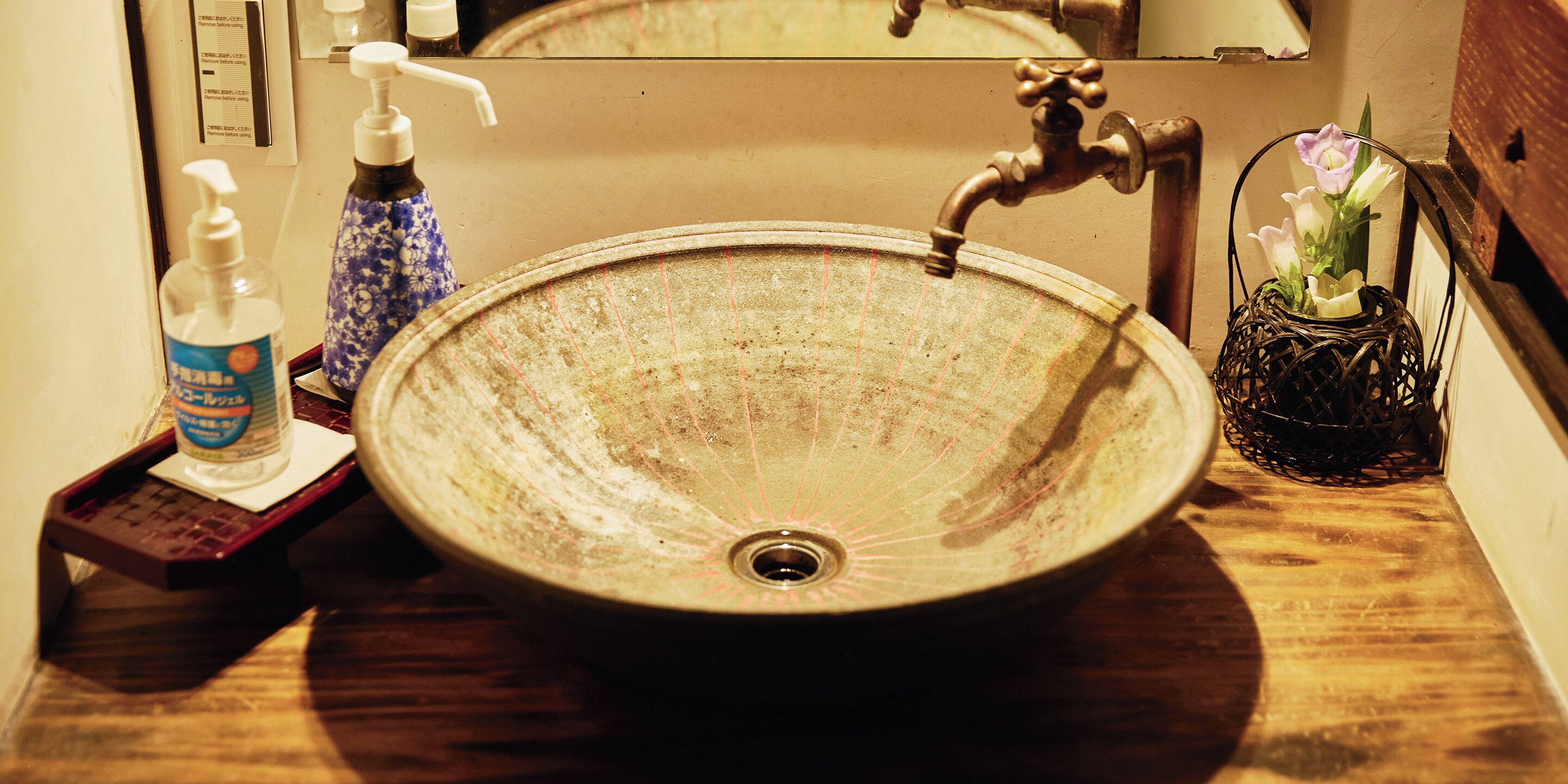 Un lavabo de cerámica redondo con un grifo de estilo antiguo, un dispensador de jabón y un jarrón decorativo con flores sobre una encimera de madera.