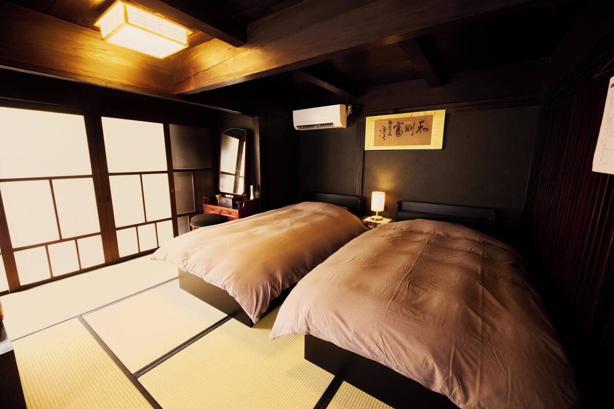 A traditional Japanese-style room with tatami flooring, two beds with matching pillows, a wooden ceiling, and a wall-mounted air conditioner.