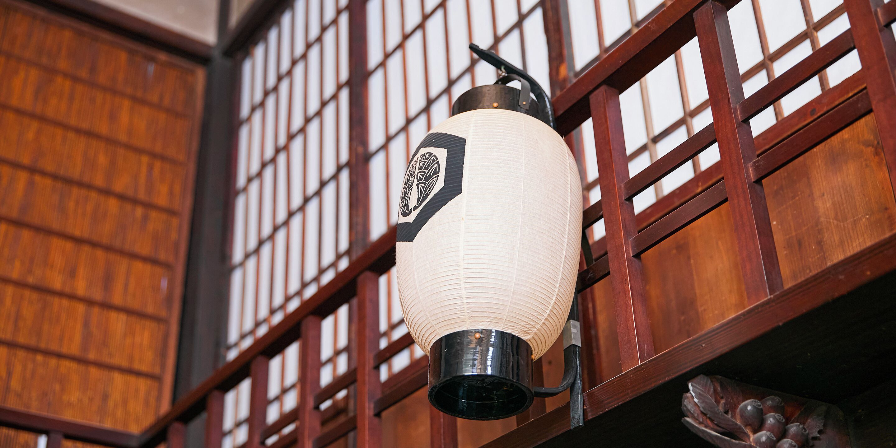 A traditional Japanese lantern hanging in front of a wooden building with a grid-like window pattern.
