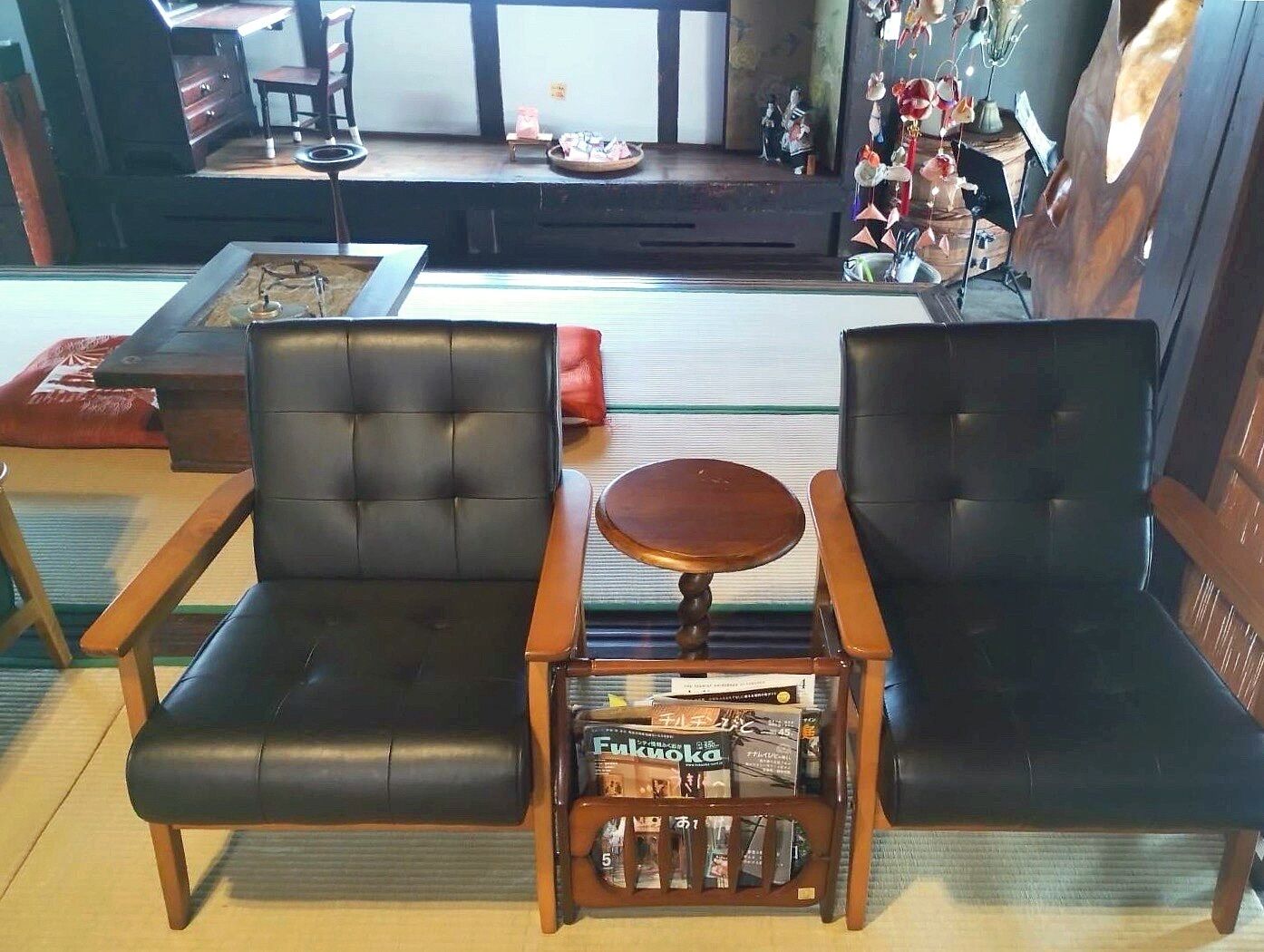 Due poltrone in pelle nera con un piccolo tavolino tra di esse, un espositore per riviste con libri e, sullo sfondo, un tavolino con un cuscino rosso.