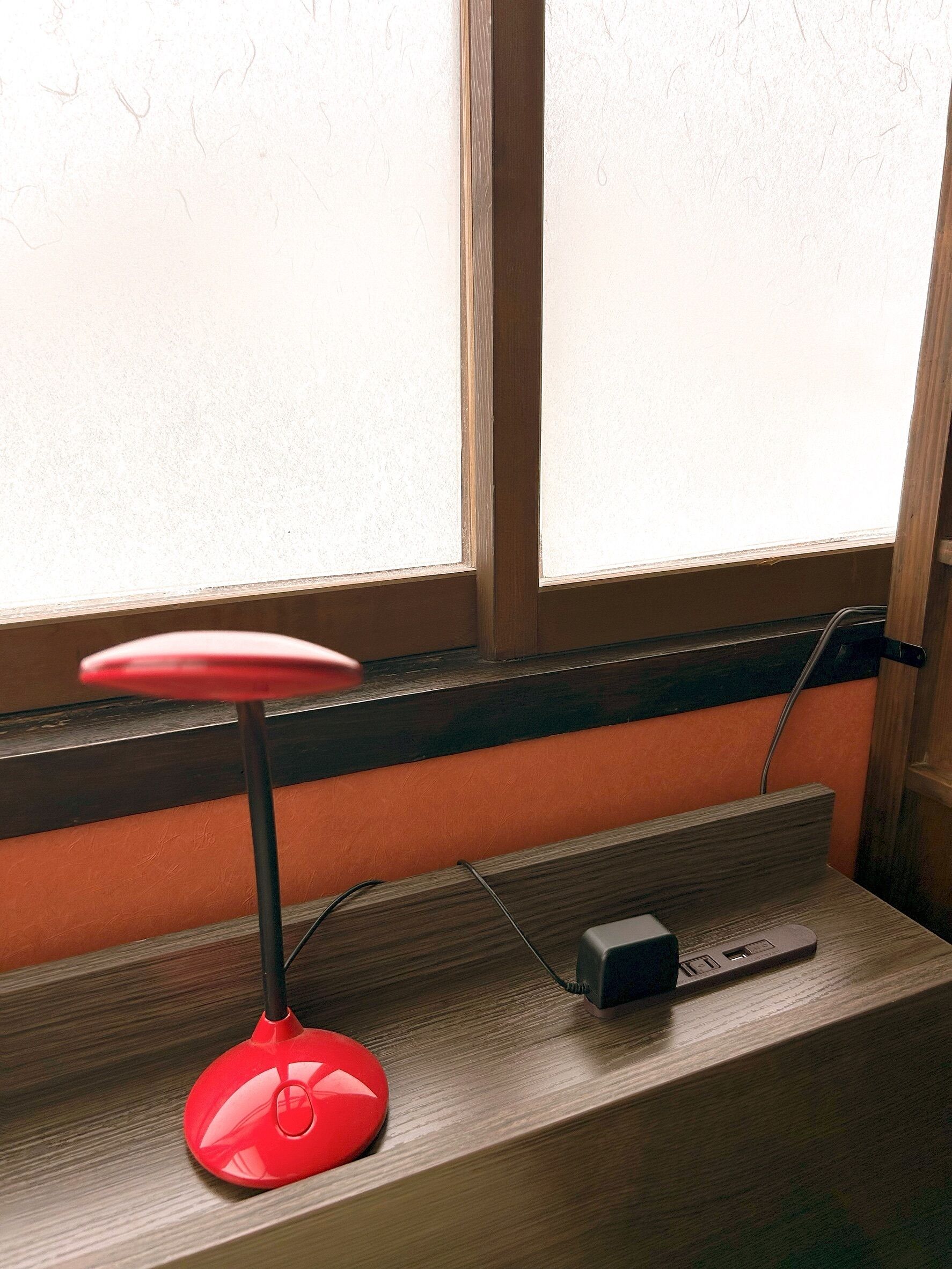 A red desk lamp on a wooden desk near a window.