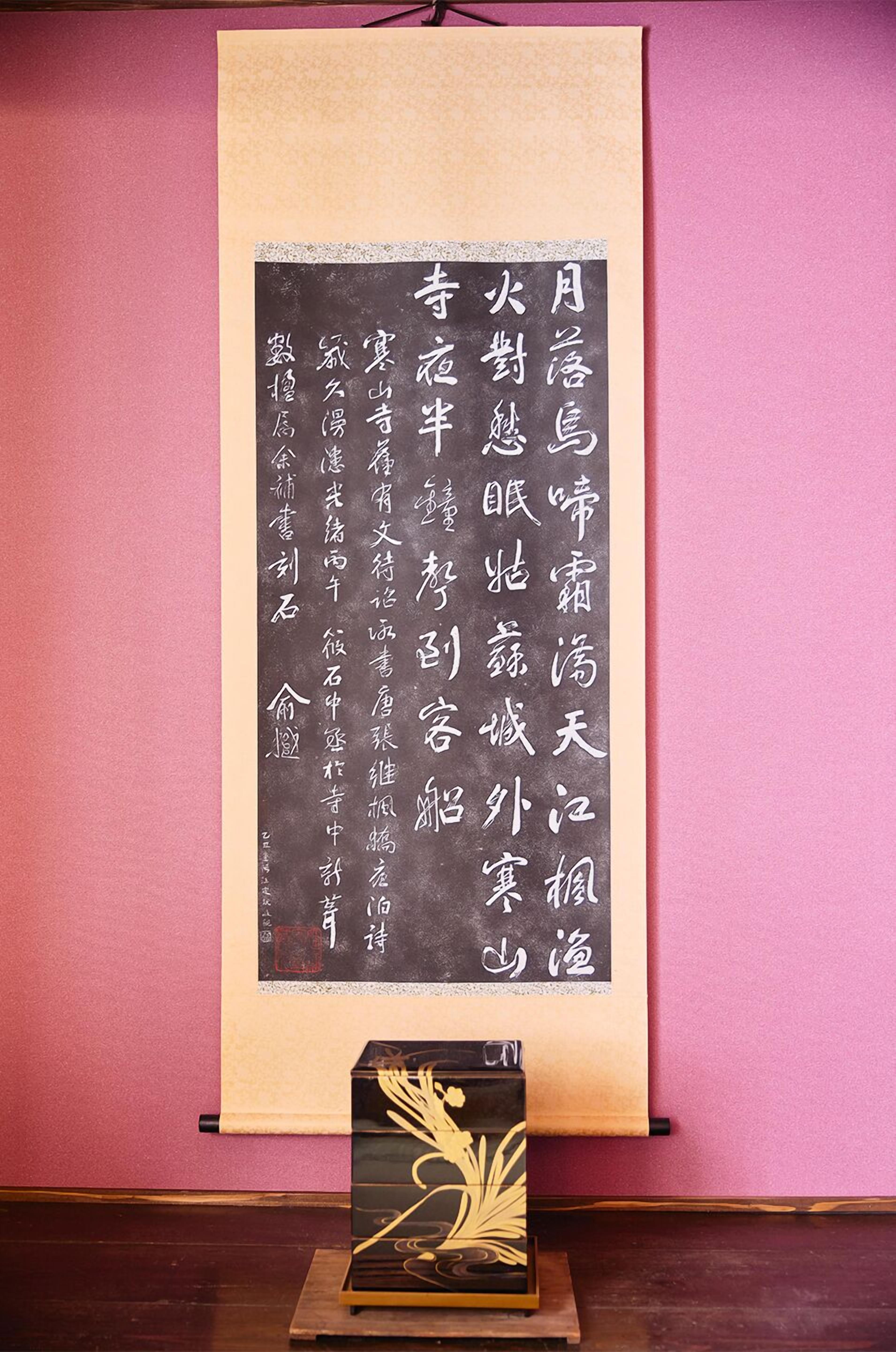 A vertical scroll with Chinese calligraphy on a beige background, displayed on a wall with a pink backdrop.