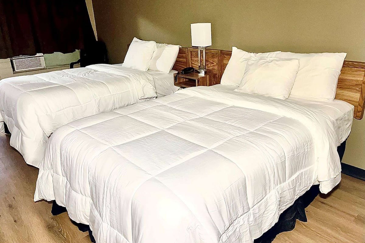 A hotel room with two beds, a bedside table with a lamp, and a wall-mounted air conditioner.