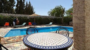 Uma área de piscina com piso de azulejos, uma mesa redonda com cadeiras e um pilar de pedra.