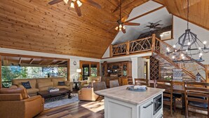 Uma sala ampla com teto de madeira, um sofá grande, uma mesa de jantar e uma cozinha com escada.