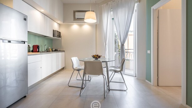 A modern kitchen with white cabinets, a stainless steel refrigerator, a microwave, and a glass dining table with four chairs. - Sleek City Oasis - City Centre (Cagliari)